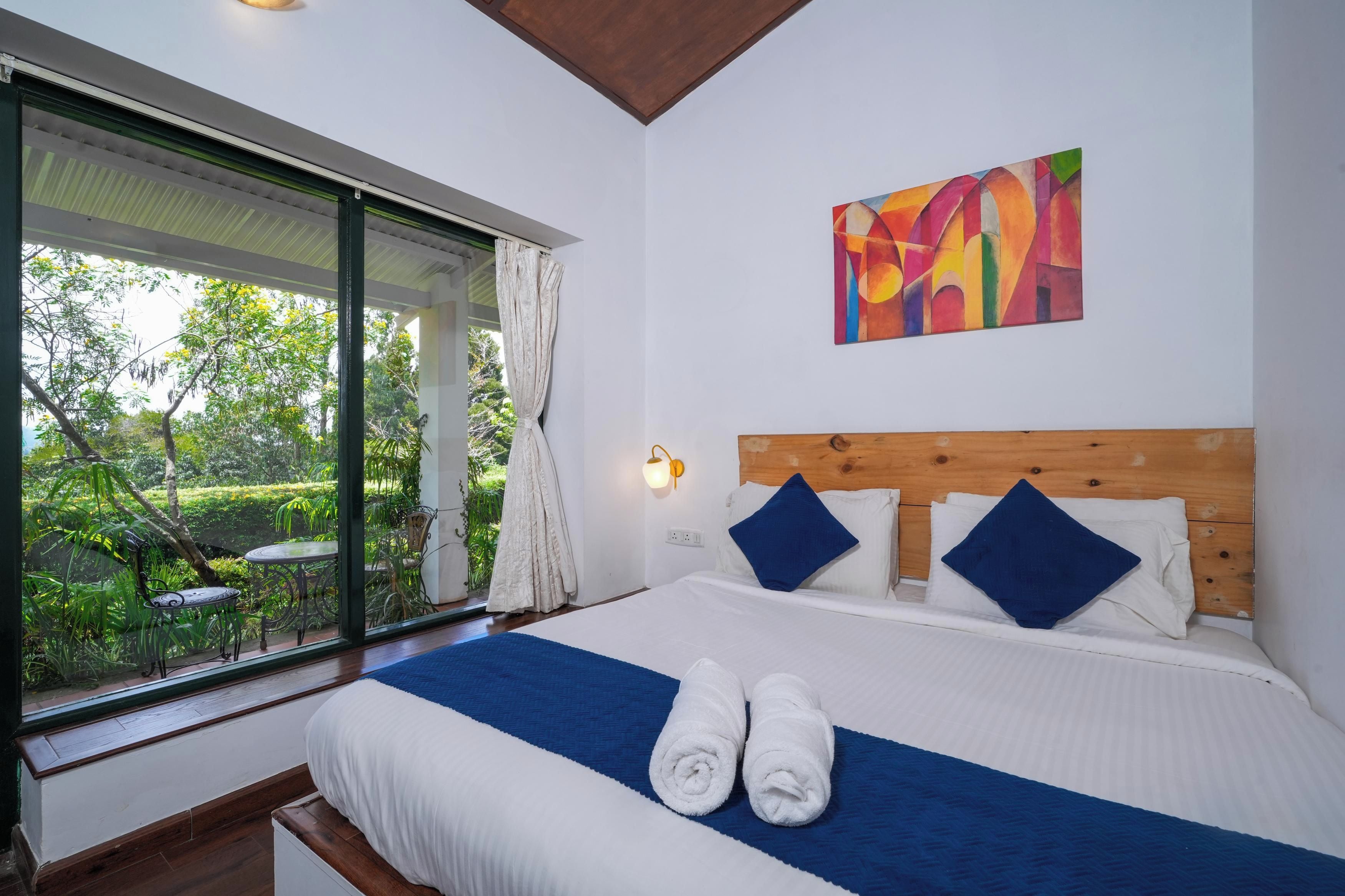 Une chambre avec un grand lit, une vue sur un jardin et une peinture abstraite aux couleurs vives accrochée au mur.