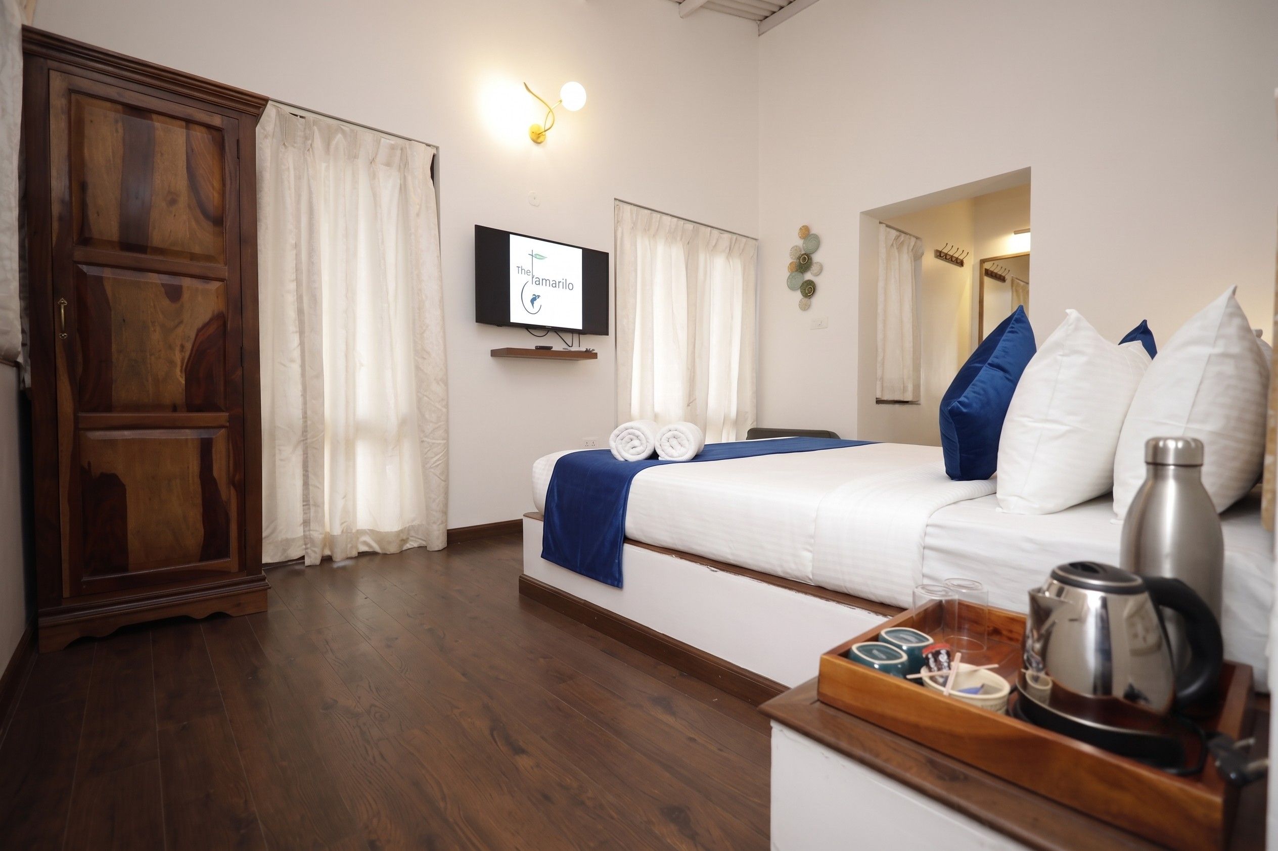 A hotel room with a wooden wardrobe, a bed with white bedding and blue accents, a flat-screen TV mounted on the wall, and a bedside table with a kettle and cups.