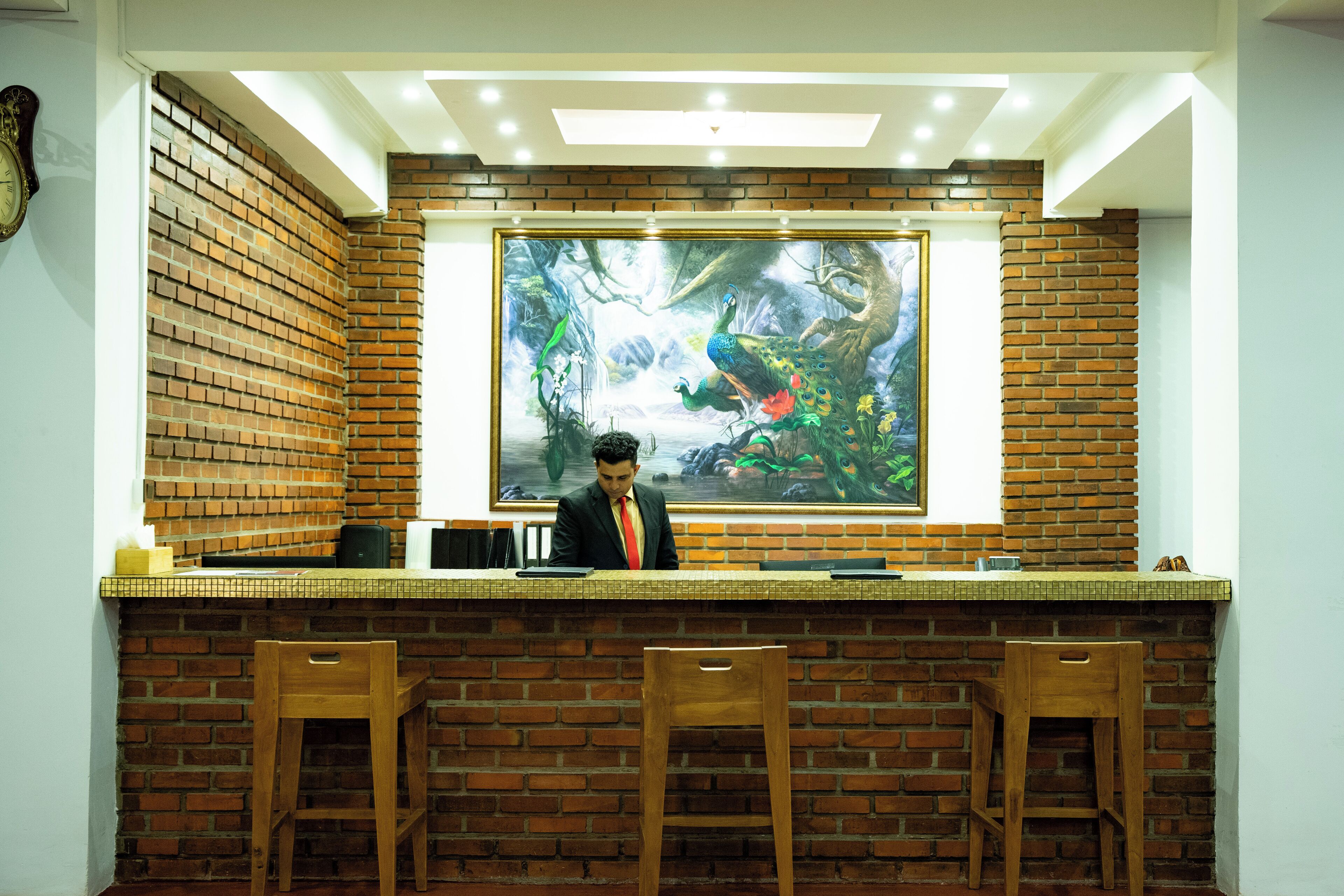 A reception area with a brick wall, a large painting of a peacock, and two wooden chairs.