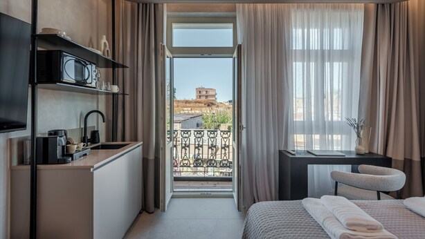 A modern hotel room with a kitchenette, a bed with neatly folded towels, a desk with a chair, and a balcony with a view of the city. - Amara Luxury Suites With Private Hot Tub (Chania)