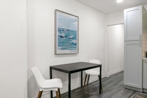 Ein kleiner Essbereich mit einem dunklen Tisch und zwei weißen Stühlen, einem gerahmten Bild an der Wand und einem weißen Schrank zur Rechten.
