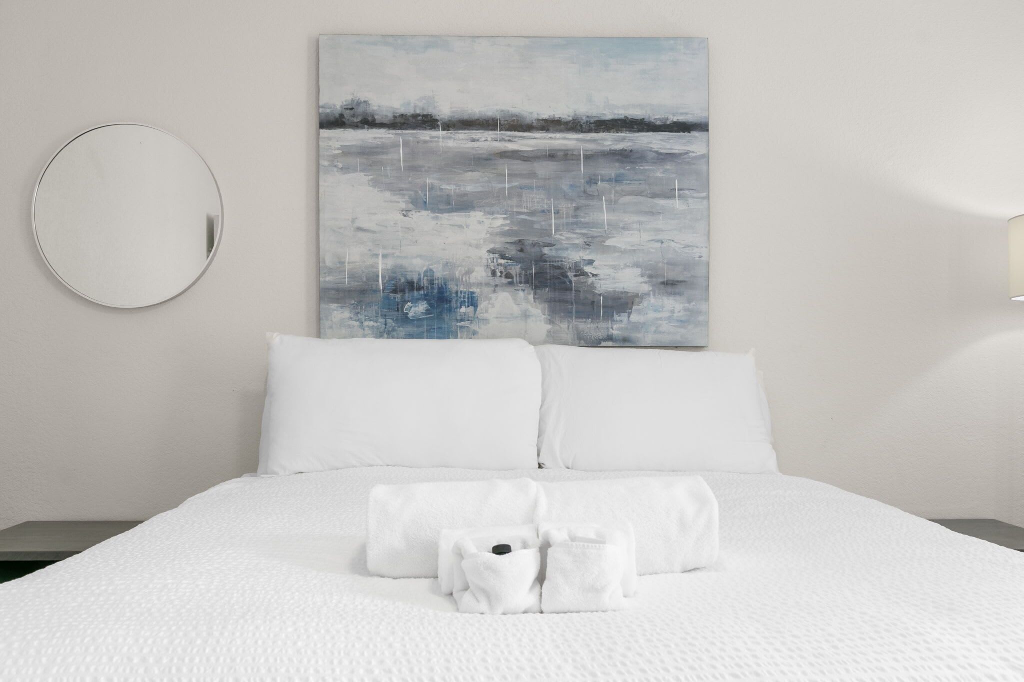 Un lit bien fait, avec du linge de lit et des serviettes blanches, un miroir rond accroché au mur et un tableau représentant un paysage.