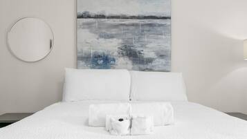 Un lit bien fait, avec du linge de lit et des serviettes blanches, un miroir rond accroché au mur et un tableau représentant un paysage.