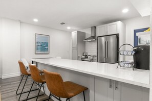 Eine moderne Küche mit weißen Schränken, Edelstahlgeräten und einer weißen Arbeitsplatte. Drei braune Stühle stehen an der Bar.