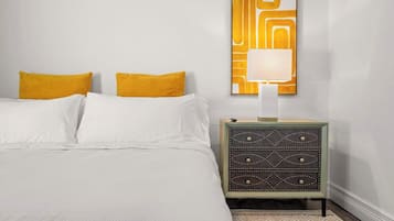 A neatly made bed with white linens and two mustard yellow pillows, a wall-mounted lamp, and a small side table with a geometric patterned drawer.