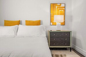 Un lit bien fait, avec du linge de lit blanc et deux coussins jaune moutarde, une lampe fixée au mur et une petite table de chevet dotée d’un tiroir à motifs géométriques.