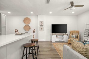 A modern kitchen with white countertops, two bar stools, a large flat-screen TV, and a woven chair.