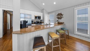 A kitchen with white cabinets, a granite countertop, a microwave, a stove, and a sink. There are three stools with cushions.