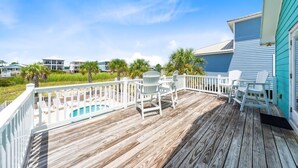 A wooden deck with white chairs and a pool, overlooking a beach with palm trees and other buildings.