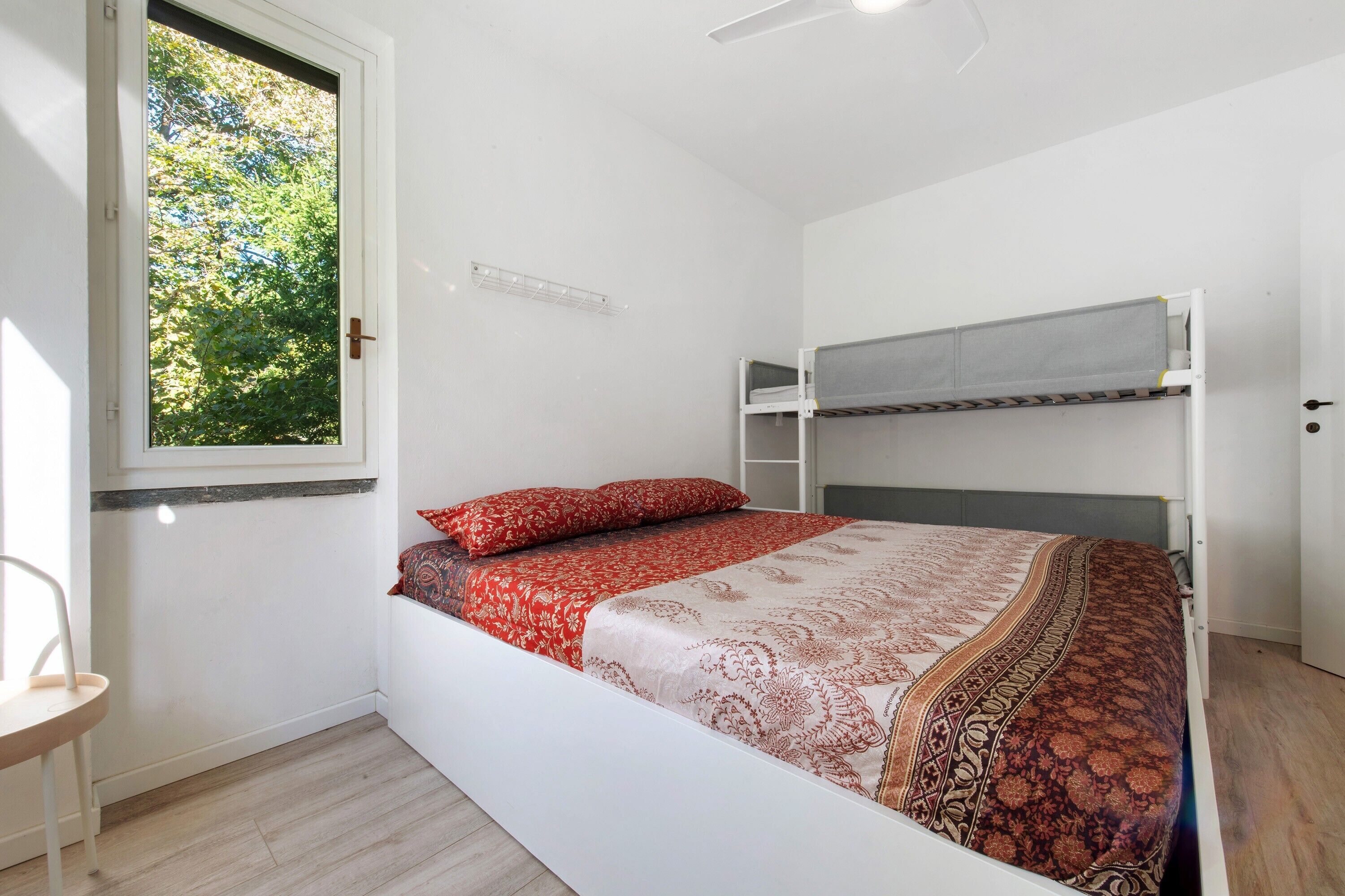 Un lit simple avec une couvre-lit à motifs, une fenêtre donnant sur des arbres et un ventilateur de plafond blanc.