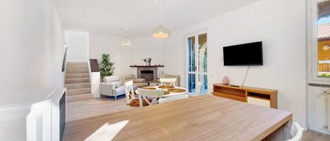 Ein modernes Wohnzimmer mit Esstisch aus Holz, weißen Sofas und einem an der Wand montierten Fernseher.