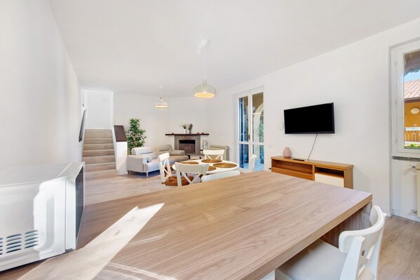 A modern living room with a wooden dining table, white sofas, and a TV mounted on the wall.