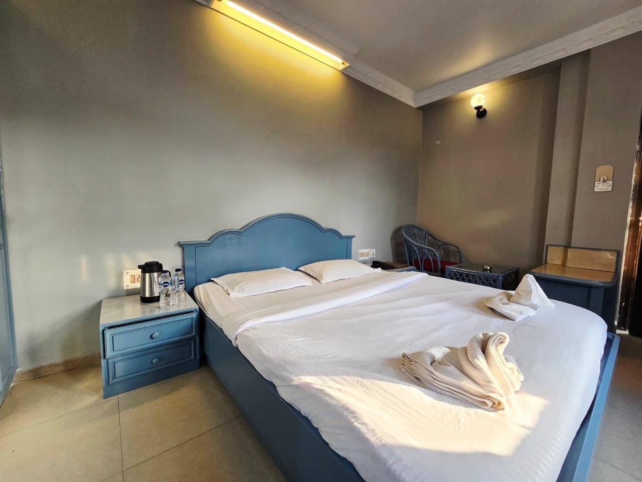 A neatly made bed with white linens and towels, a blue headboard, and a nightstand with a coffee maker and bottles.