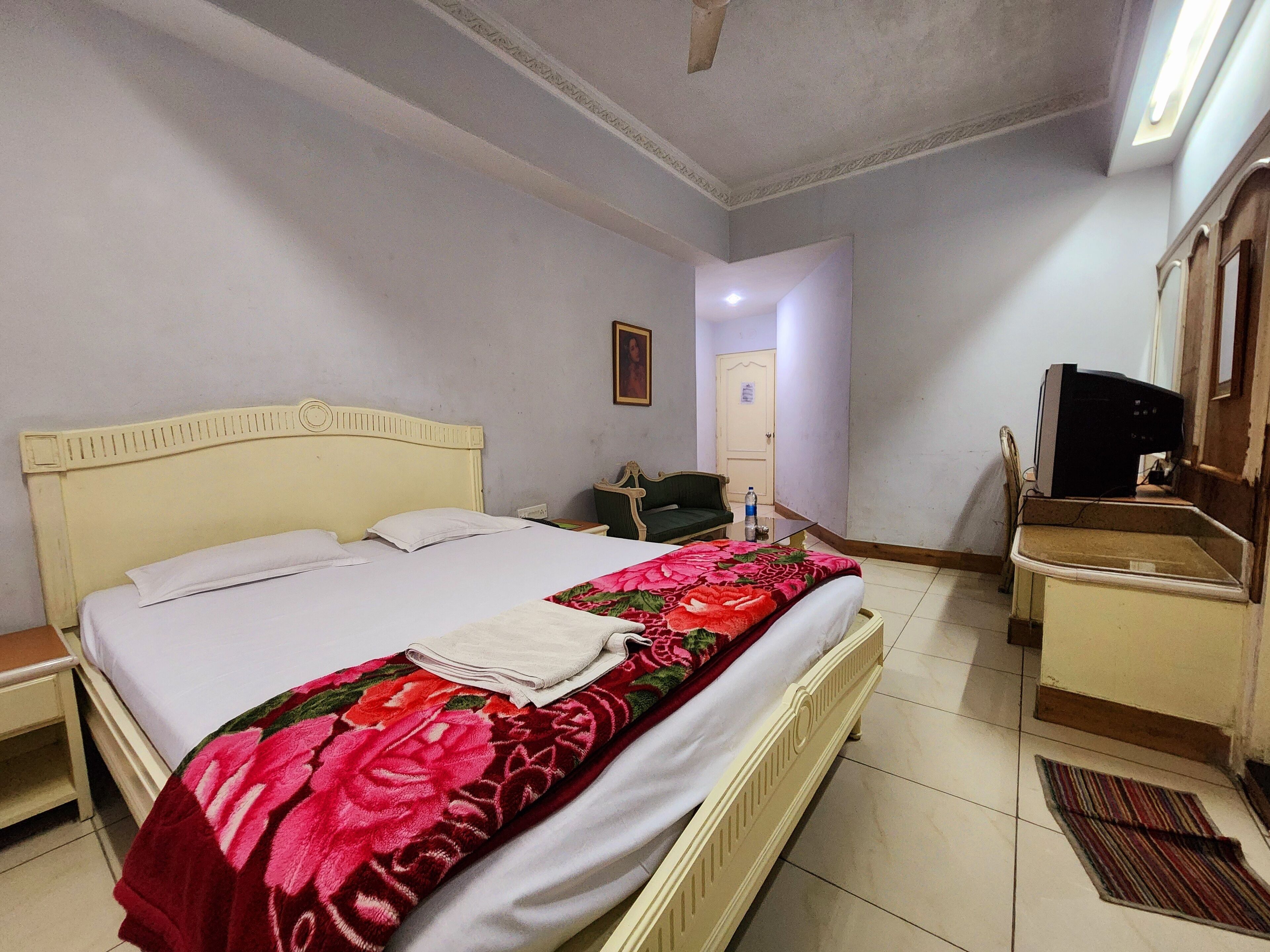 A hotel room with a bed, a nightstand, a dresser, a television, and a door leading to another room.