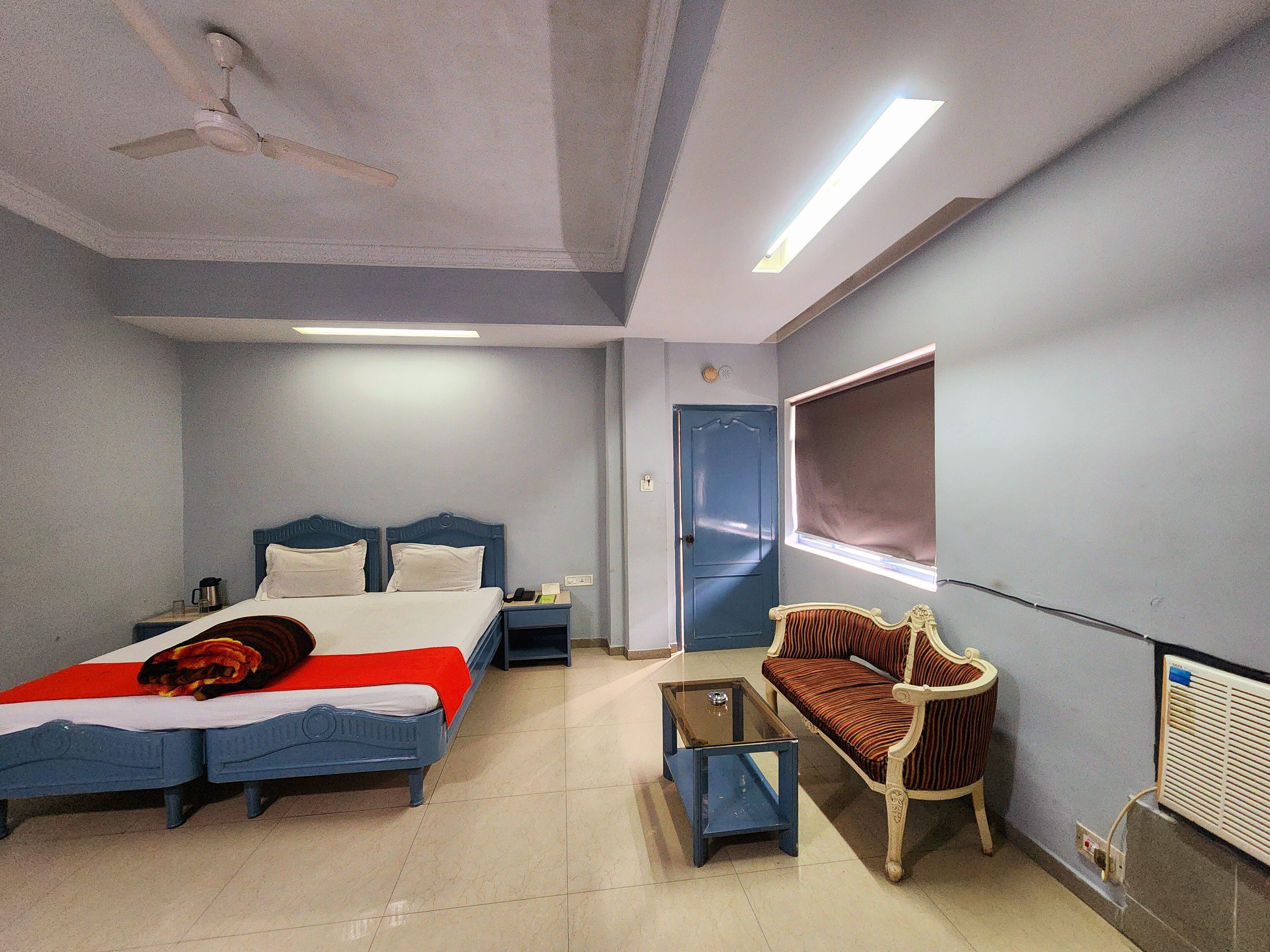 A hotel room with two beds, a sofa, a small table, a ceiling fan, and a window with blinds.