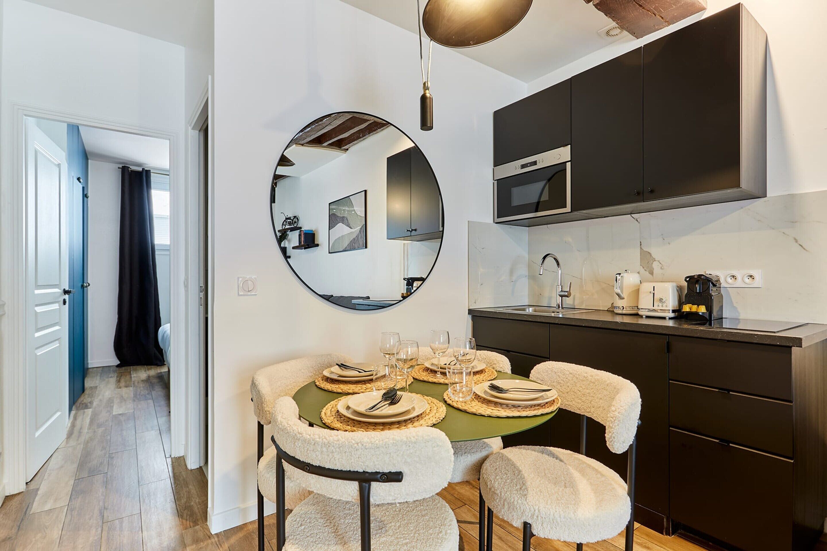 A compact kitchen with dark cabinets, a round mirror, and a dining table set for six with chairs.