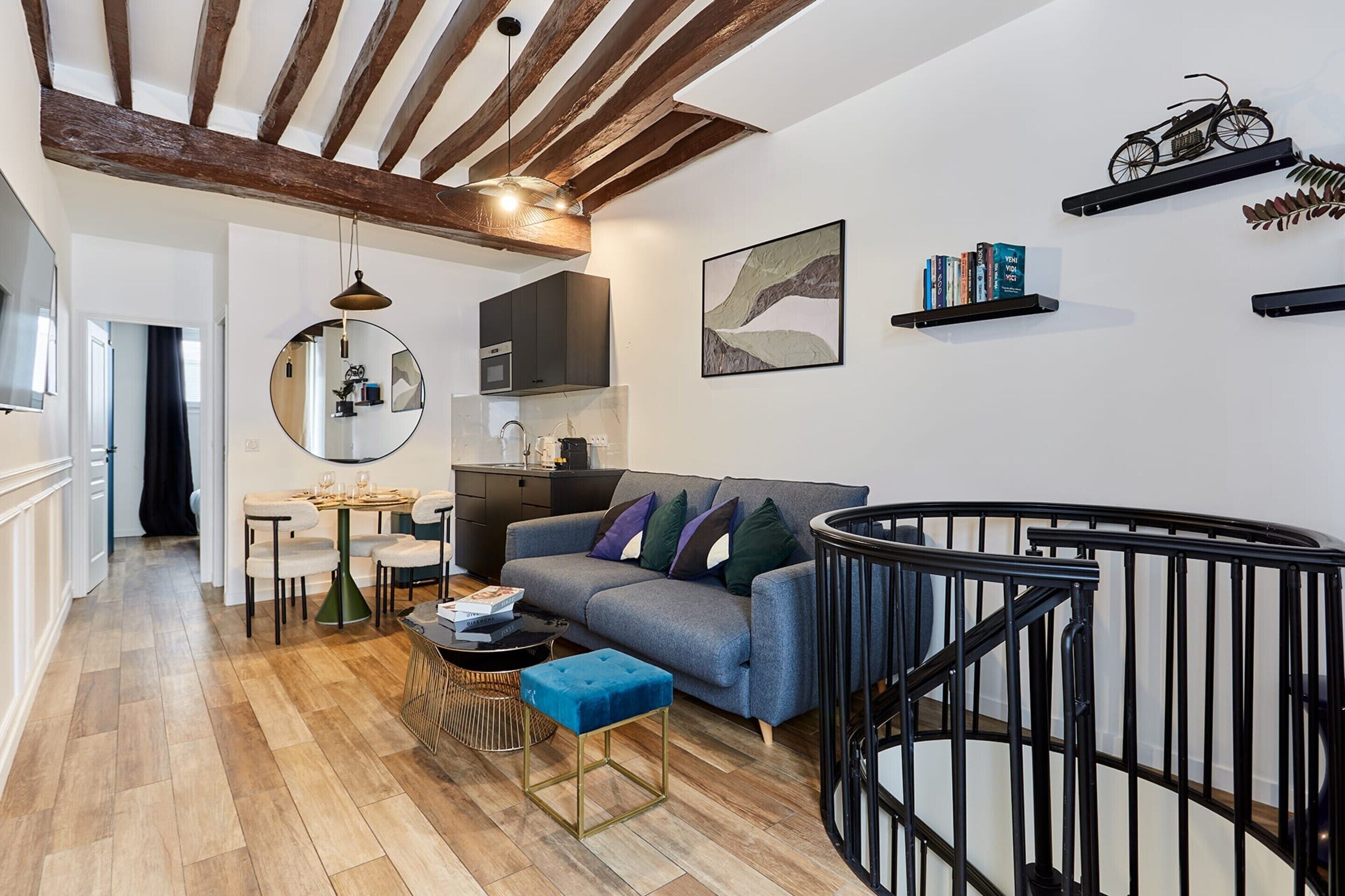 Una sala moderna con un sofá gris, un comedor con una mesa redonda y una escalera con barandilla negra.