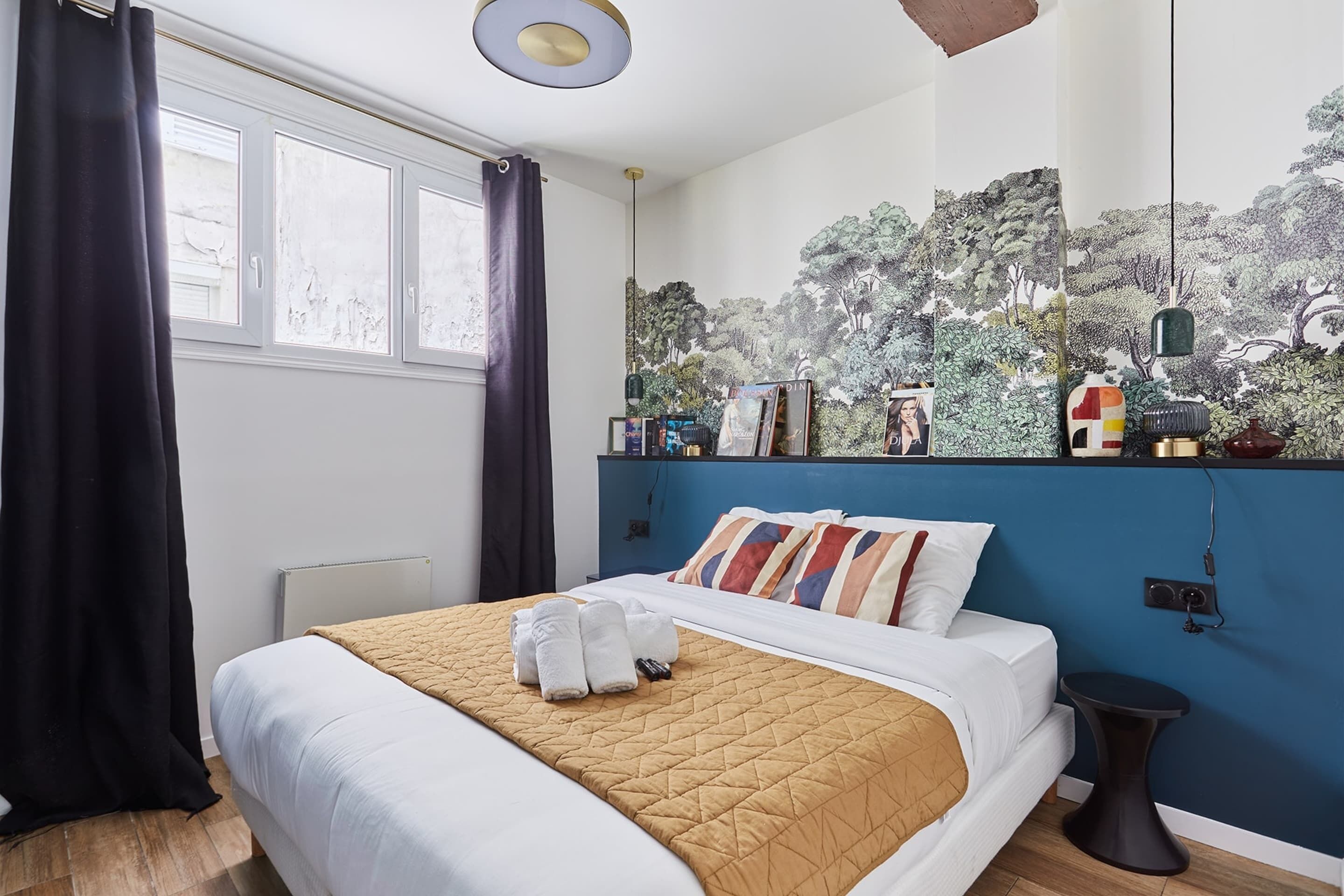 A hotel room with a bed, a window with curtains, a wall mural, and a bedside table.