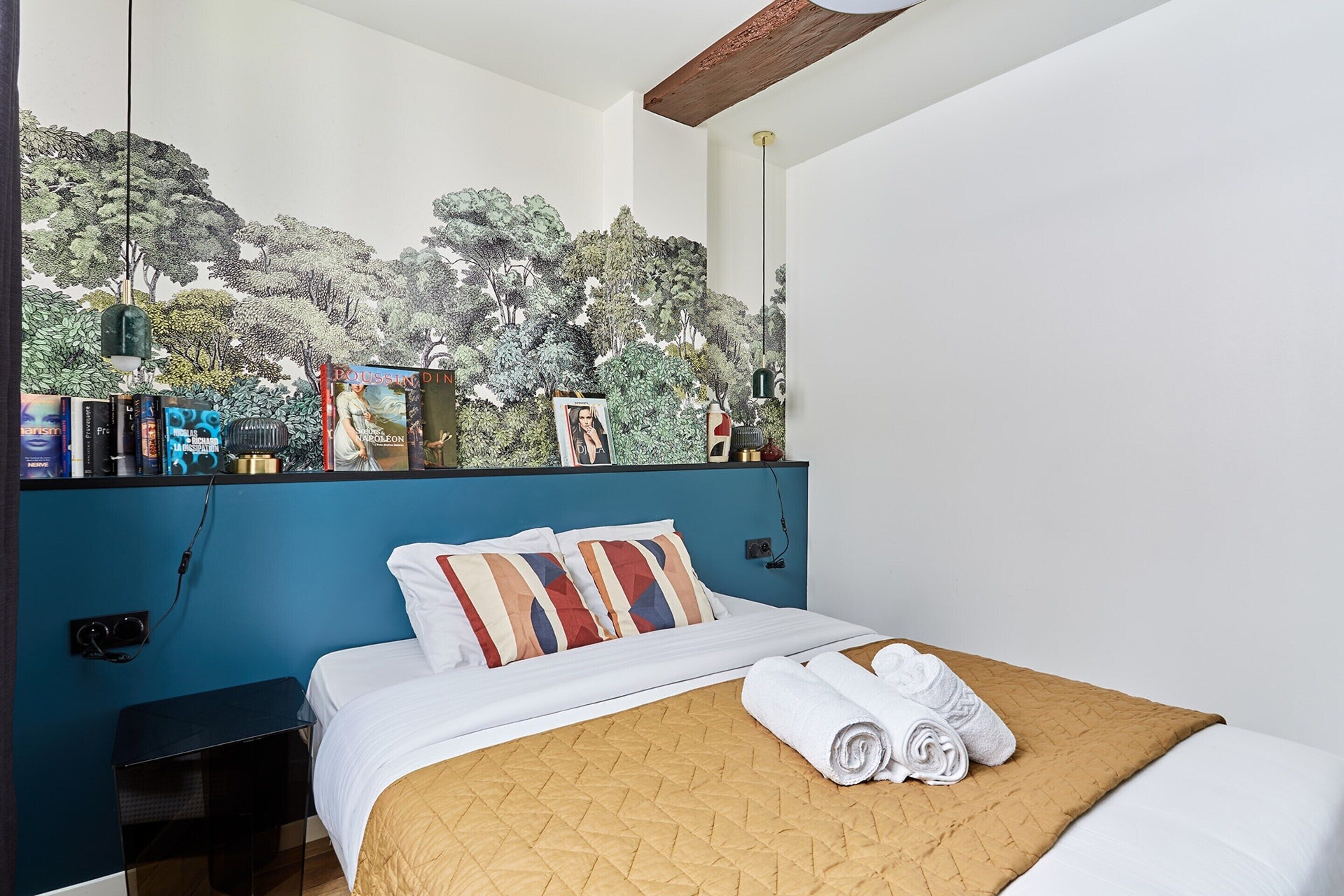 A hotel room with a bed, a nightstand, a bookshelf, and a mural on the wall.