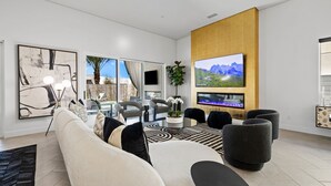 A modern living room with a sofa, coffee table, and chairs. There is a TV mounted on the wall and a fireplace below it. - Casa De Amore (Palm Springs)