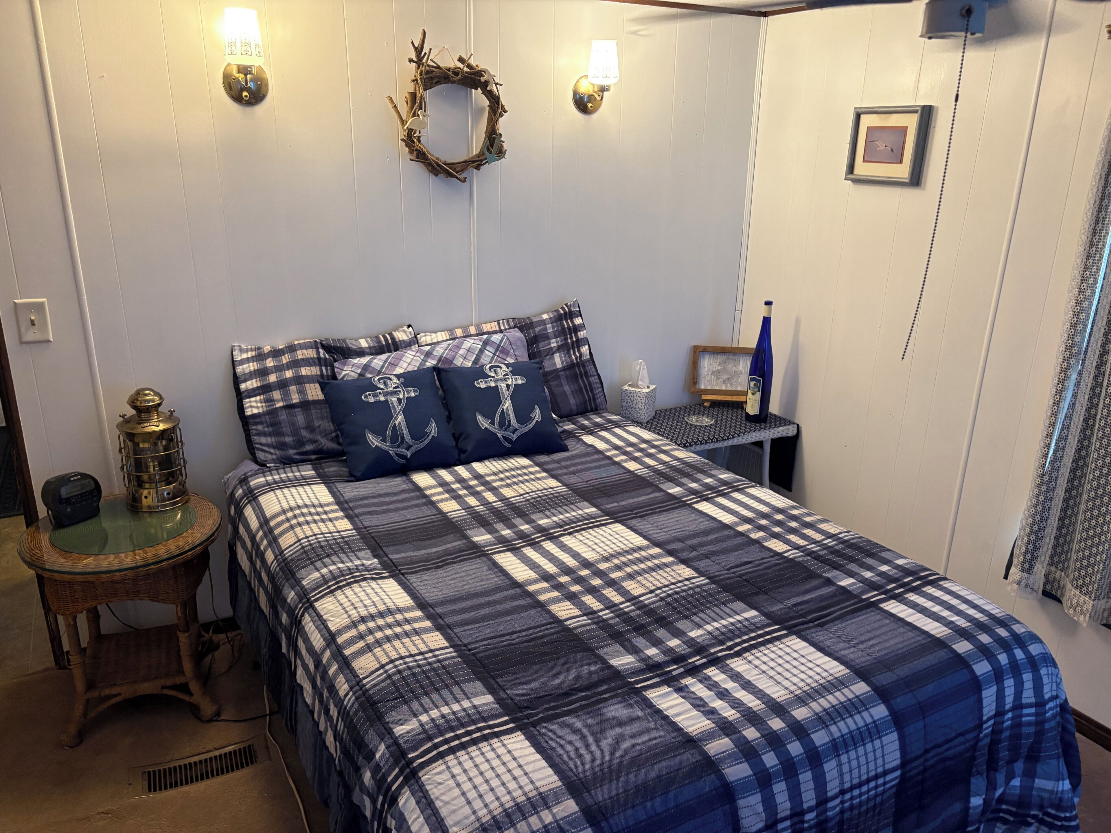 A bedroom with a plaid bedspread, a nightstand with a lamp, a wall-mounted light fixture, and a framed picture.
