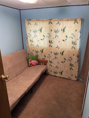 Une petite chambre accueillante, avec un lit simple, une table de chevet en bois et un rideau orné d’un motif floral.