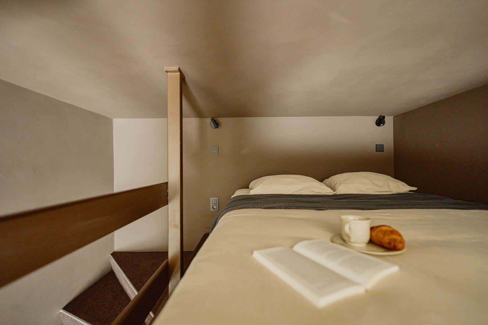 Une petite chambre avec un lit simple, une tête de lit en bois, une table de chevet avec une tasse et une pâtisserie, et un livre posé sur le lit.
