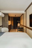 A hotel room with a bed, a desk with a chair, a hanging lamp, and a wardrobe.