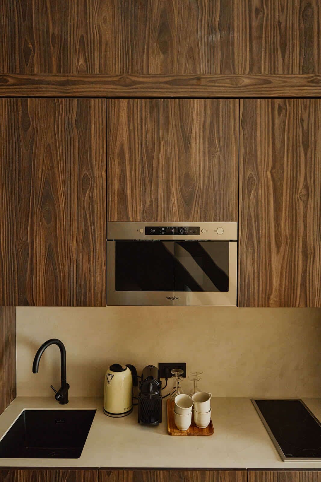 Eine moderne Küche mit einem eingebauten Edelstahlbackofen, einem schwarzen Wasserhahn, einem Wasserkocher und einer Kaffeemaschine auf der Arbeitsfläche.