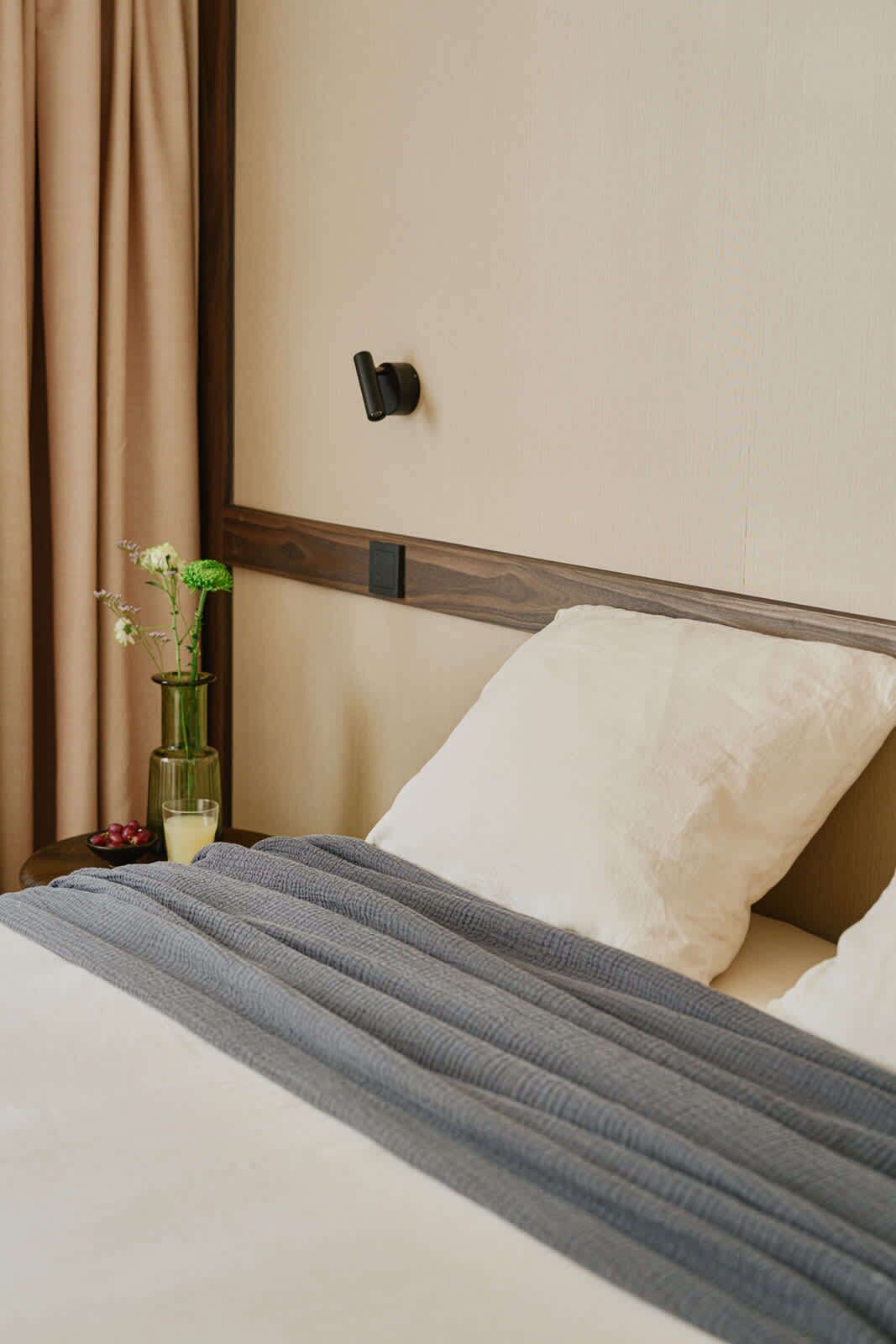Un lit bien fait, recouvert d’une couverture à rayures grises, avec un oreiller blanc et un vase de fleurs posé sur une table de chevet.