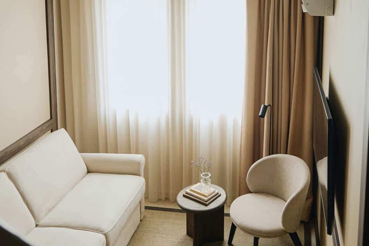 Ein Hotelzimmer mit einer Couch, einem Sessel, einem kleinen Tisch mit Buch und Vase sowie einem großen Fenster mit Vorhängen.