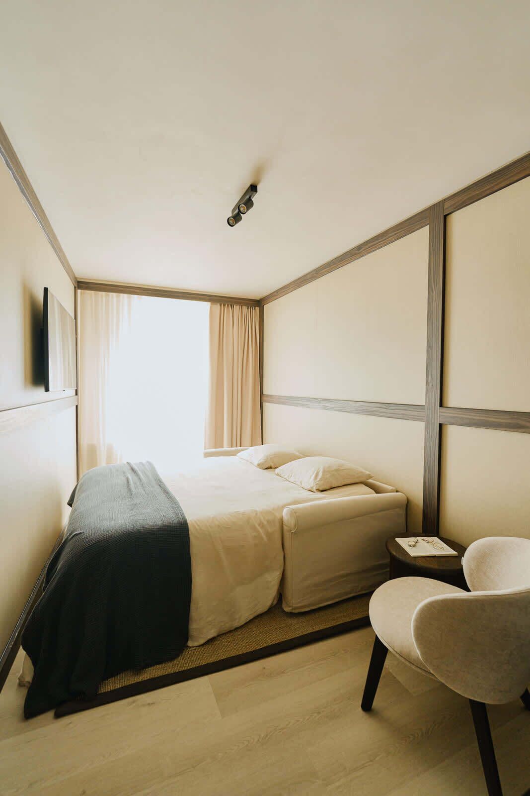 Habitación de hotel con una cama, una silla, una mesita y un ventanal con cortinas.