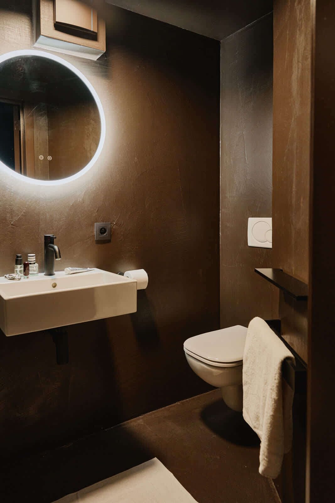 A modern bathroom with a round mirror, a sink with toiletries, and a toilet.
