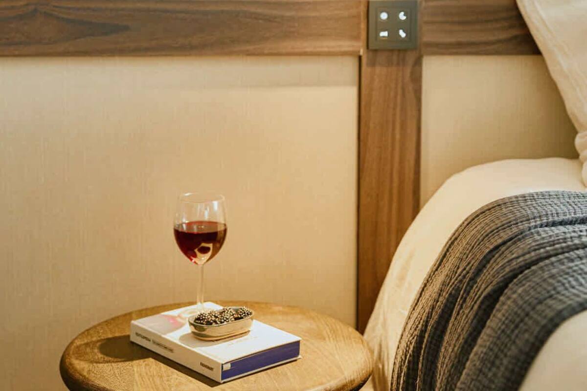 Ein Nachttisch aus Holz mit einem Buch und einem Glas Rotwein, eine Holzvertäfelung an der Wand und ein Bett mit gestreifter Decke.
