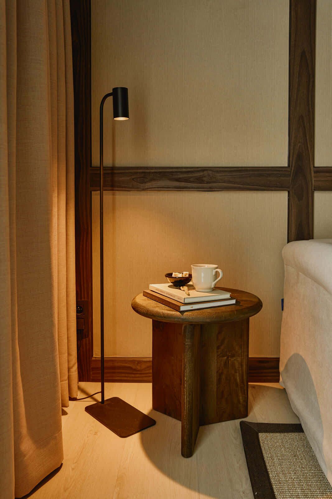 A cozy hotel room with a wooden side table, a floor lamp, a bed, and a small rug.