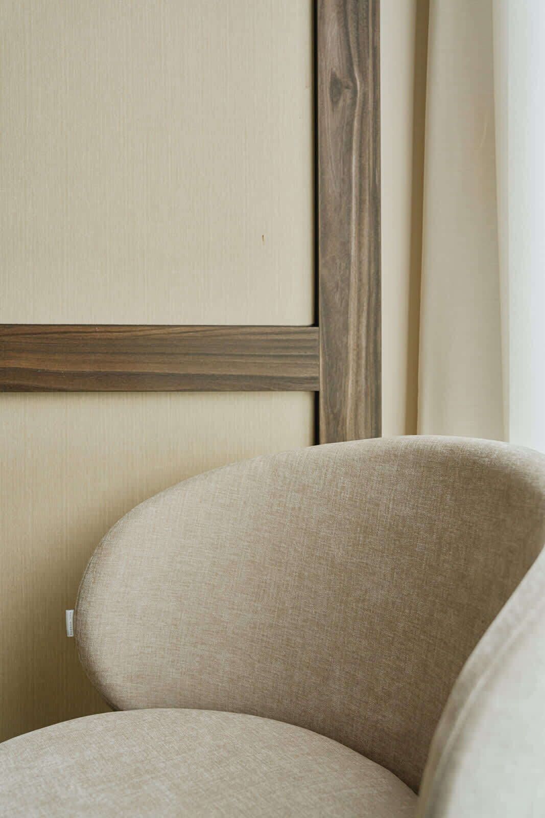 A corner of a room with a beige wall, a wooden trim, and a beige upholstered chair.