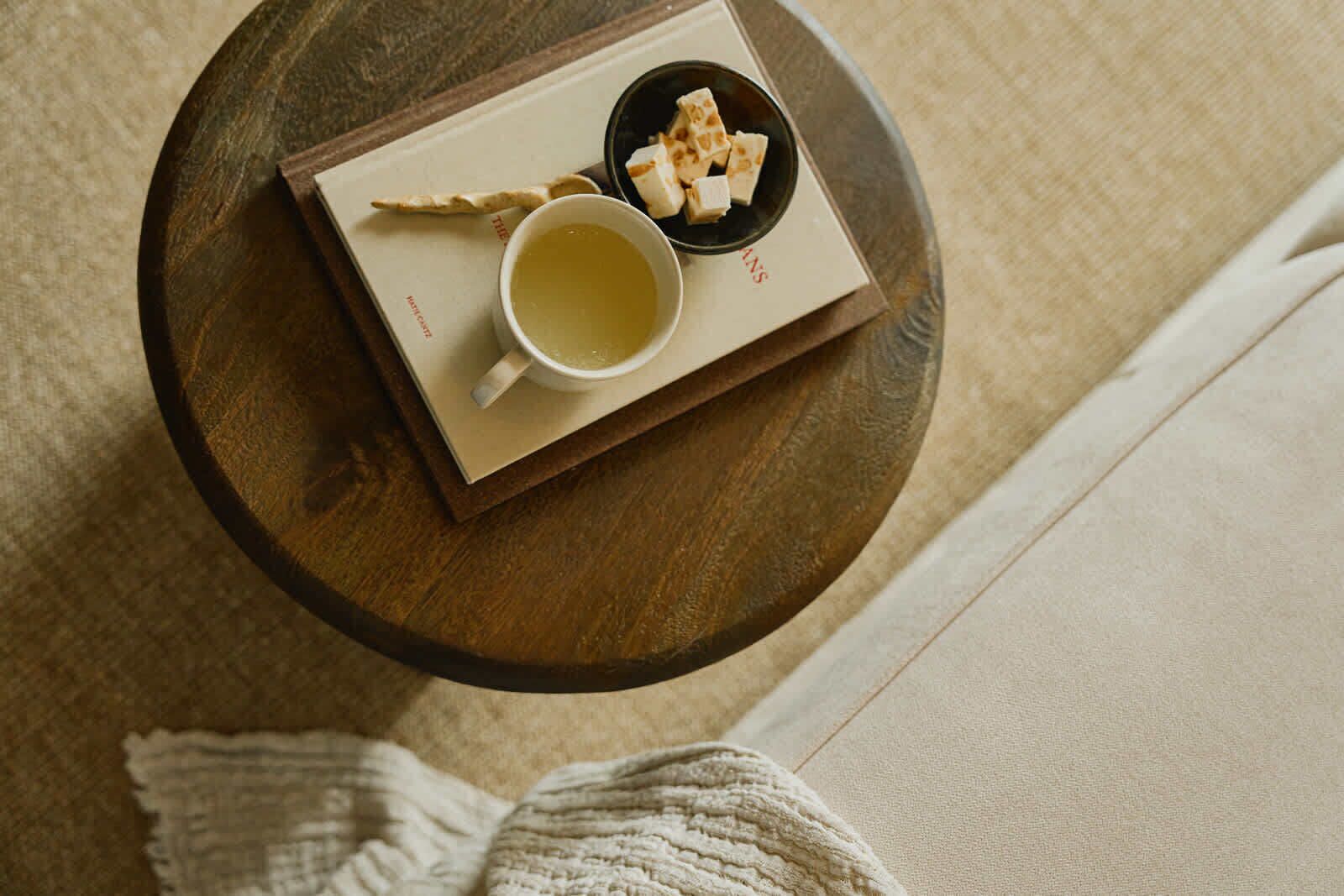 A round wooden table with a white mug, a small bowl of cubes, and a book.