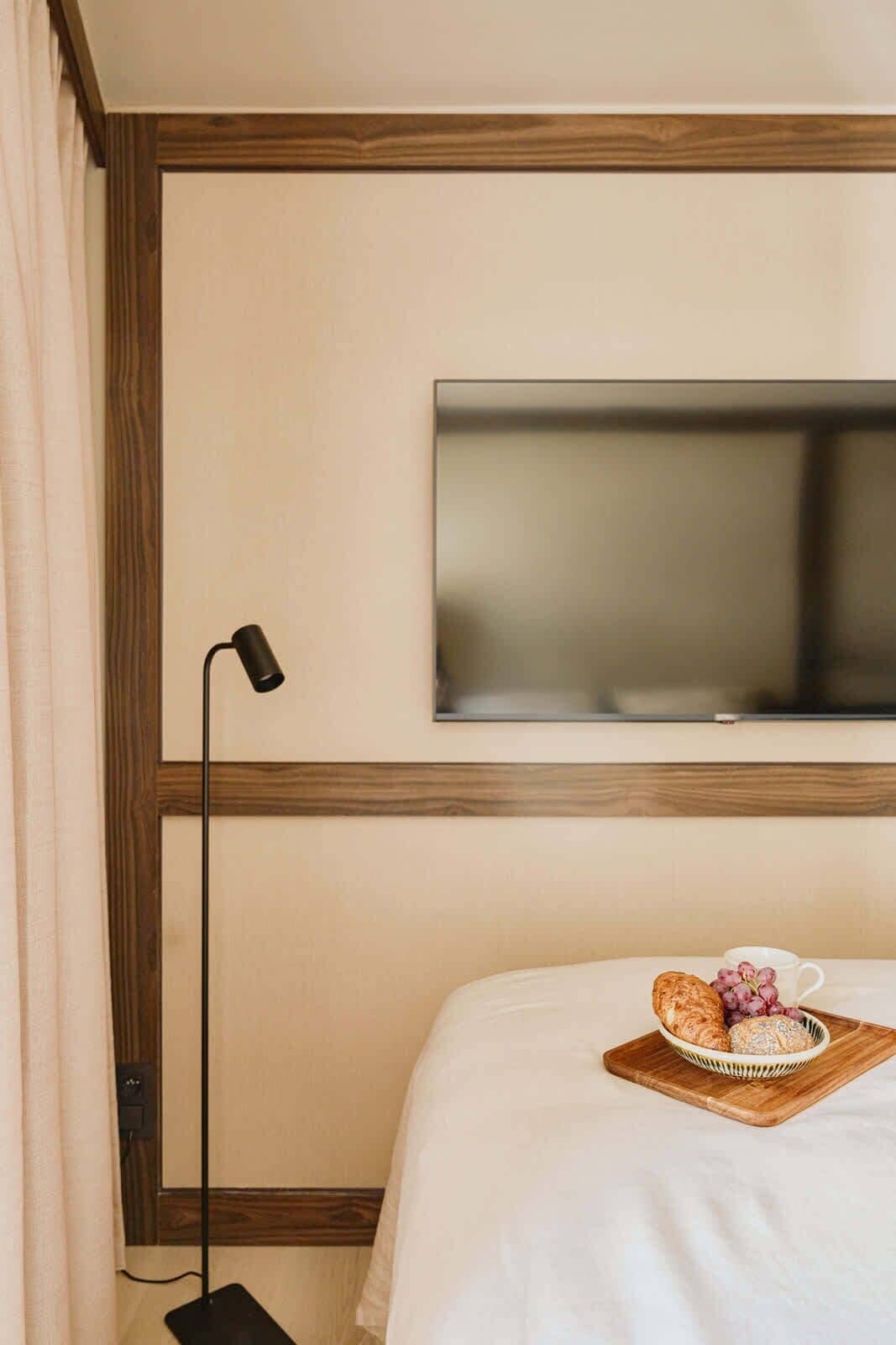 A hotel room with a bed, a TV mounted on the wall, a bedside table with a tray of pastries, and a floor lamp.