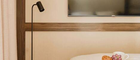 A hotel room with a bed, a TV mounted on the wall, a bedside table with a tray of pastries, and a floor lamp.