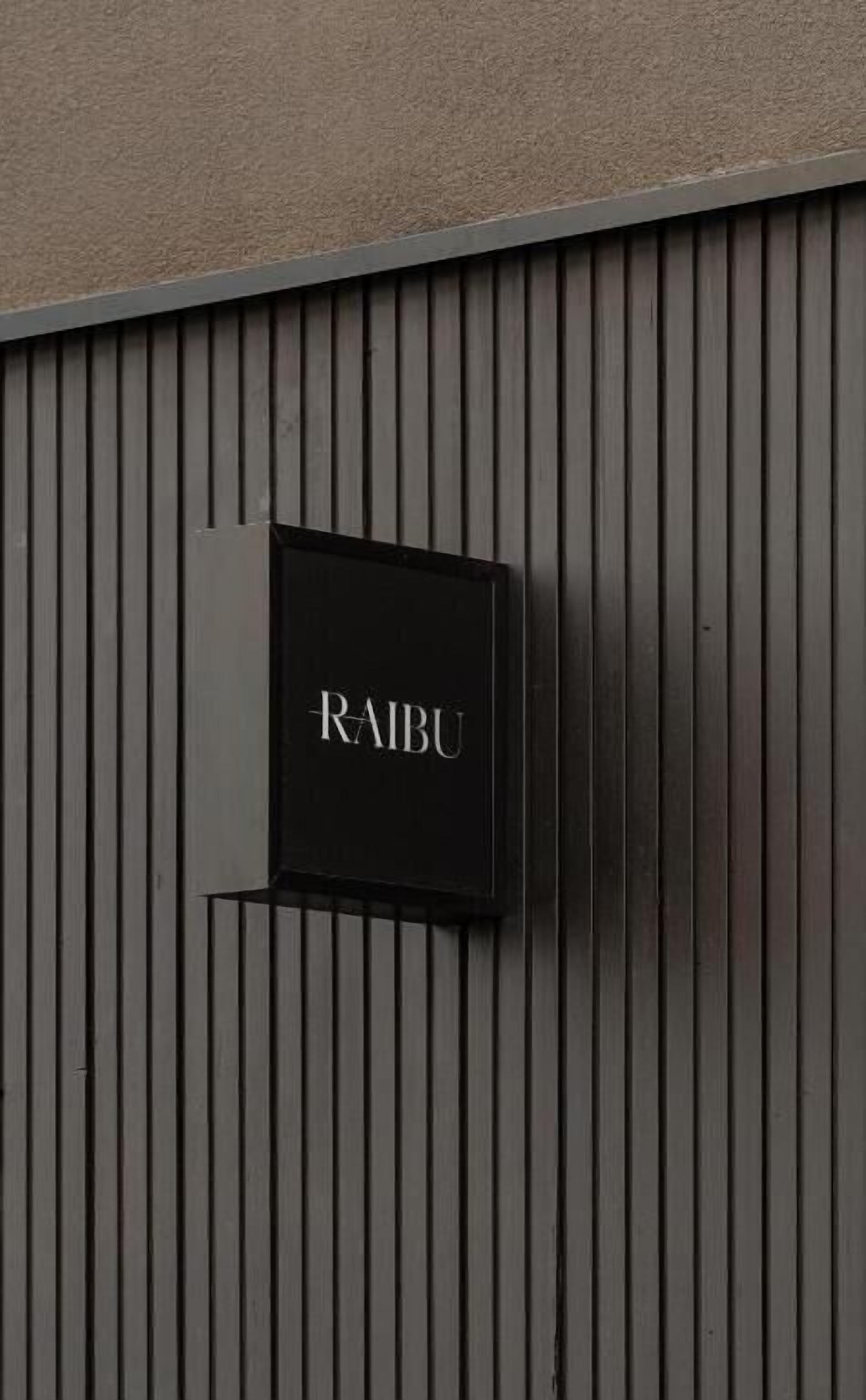 Un letrero negro con la palabra "RAIBU" sobre una pared texturizada con líneas verticales.