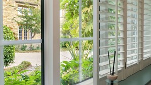 A room with a large window, a white interior, and a view of a garden and a building outside. - Beautiful 2BD Cottage in the Cotswolds (Churchill)