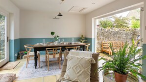 A dining area with a table set for four, surrounded by wooden chairs, a potted plant, and a stone wall visible through an open door. - Beautiful 2BD Cottage in the Cotswolds (Churchill)