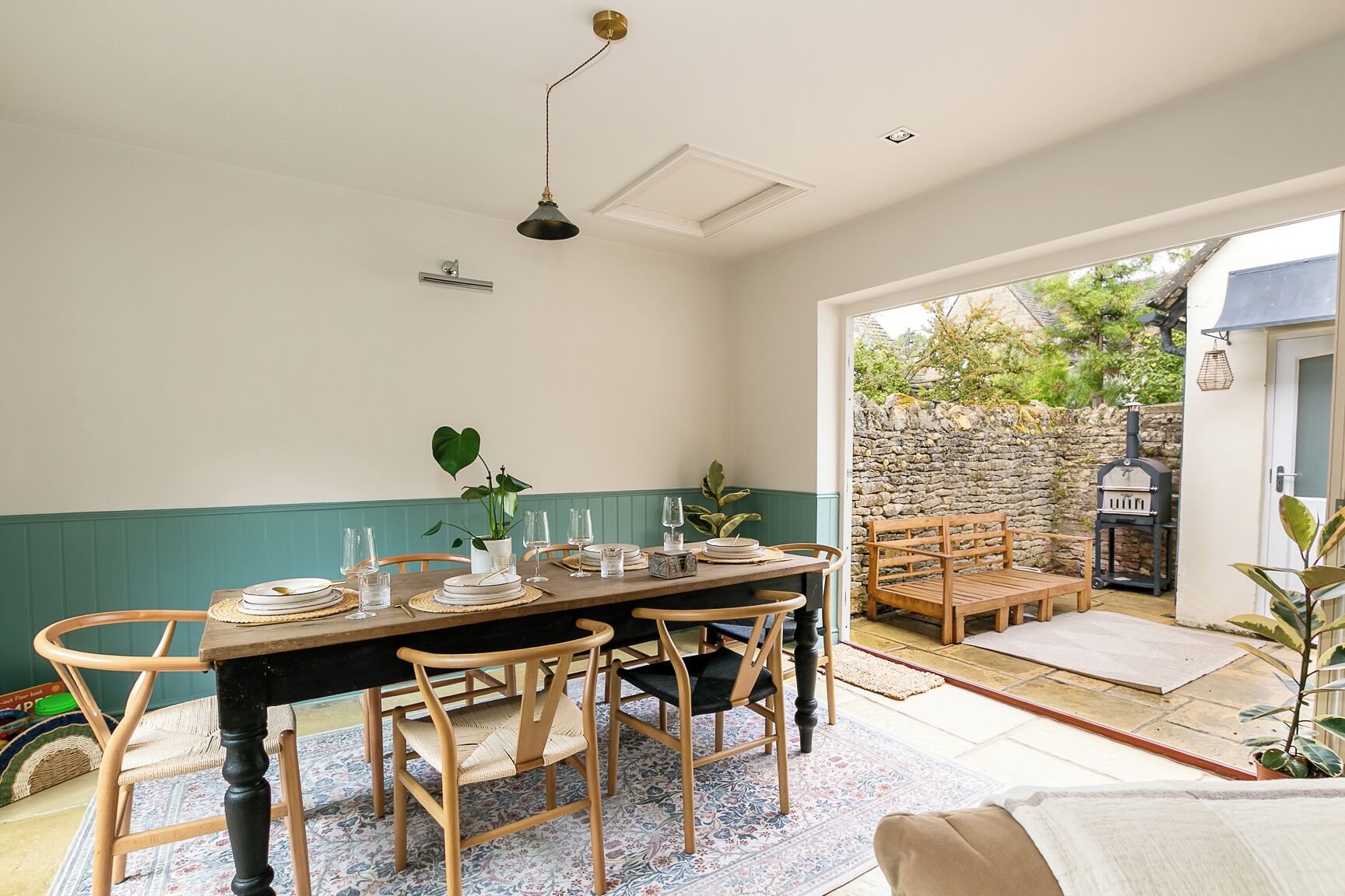 Un coin repas avec une table en bois dressée pour six personnes, entourée de chaises en bois. On aperçoit un mur en pierre et un banc en bois à l’arrière-plan.