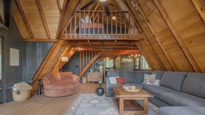 A cozy loft-style living room with a high ceiling, wooden beams, a plush sofa, a coffee table, and a staircase leading to an upper level.