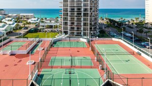 Ein mehrstufiger Tennisplatzkomplex mit Blick auf den Ozean und einem hohen Gebäude im Hintergrund.