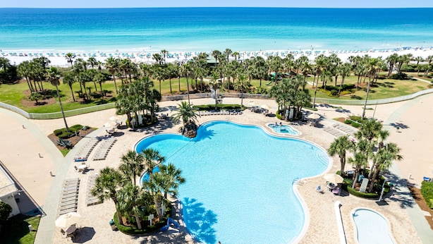 Ein großer, unregelmäßig geformter Swimmingpool, umgeben von Palmen und Liegestühlen, mit einem Strand und dem Meer im Hintergrund.