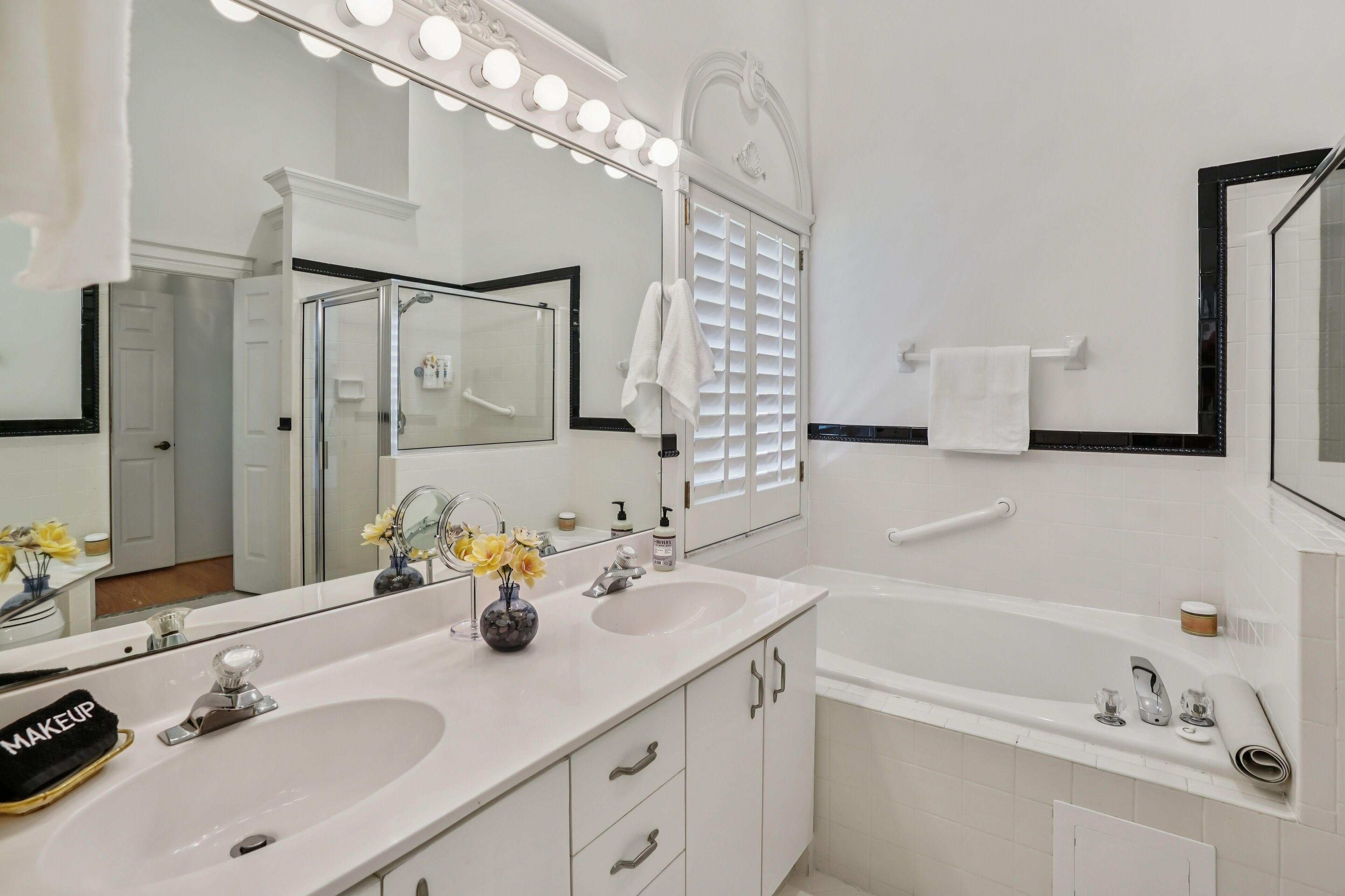 A bathroom with two sinks, a large mirror with lights, a bathtub, and a towel rack.