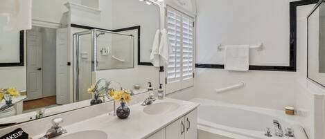 A bathroom with two sinks, a large mirror with lights, a bathtub, and a towel rack.