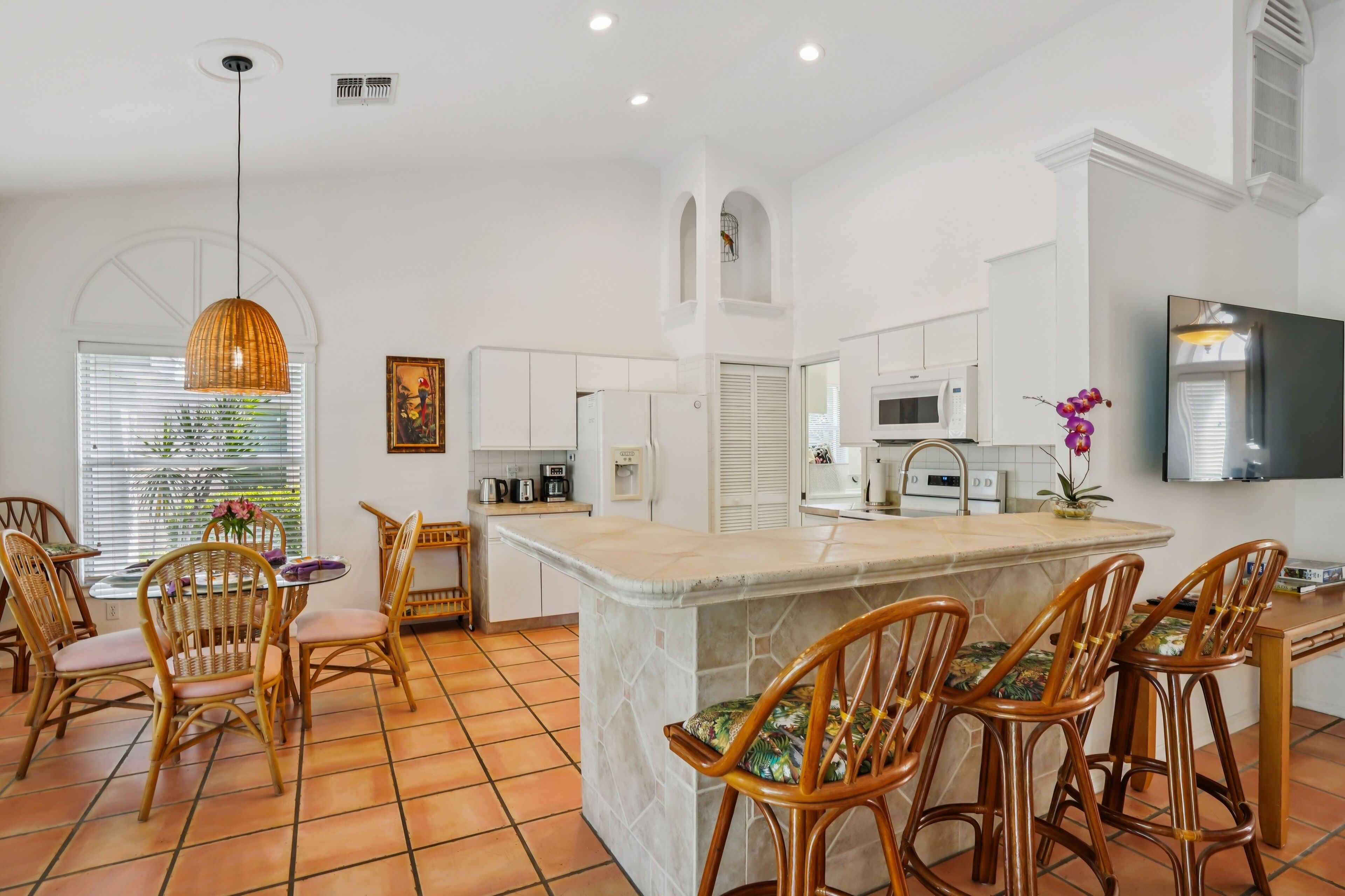 A modern kitchen with white cabinets, a central island with bar stools, a hanging pendant light, and a flat-screen TV mounted on the wall.