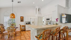 Une cuisine moderne avec des meubles blancs, un îlot central doté de tabourets de bar, un luminaire suspendu et un téléviseur à écran plat fixé au mur.